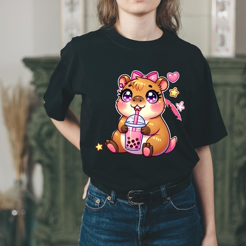 以下が含まれることがあります： 黒いTシャツに、ピンクのリボンをつけたハムスターがバブルティーを飲んでいる漫画のイラスト。星とハートが描かれています。カラフルで遊び心のあるデザインです。