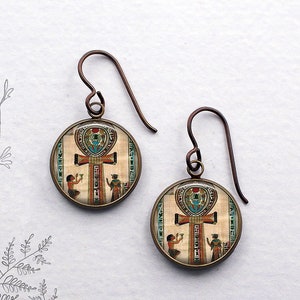 May include: A pair of bronze-toned earrings with a round glass pendant featuring an image of the Egyptian Ankh symbol. The image is surrounded by a decorative border and includes two figures on either side of the symbol.