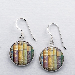 May include: Silver dangle earrings with round glass charms featuring a vintage book stack design. The books have spines with gold lettering and colorful illustrations.