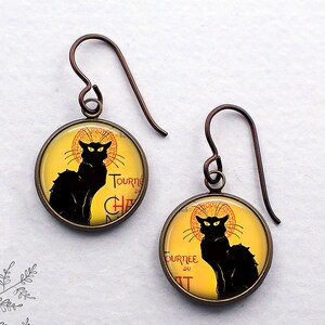May include: A pair of round earrings featuring a black cat silhouette on a yellow background, with text in red and black. The earrings have bronze-colored ear wires.