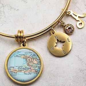 May include: Gold charm bracelet with a compass charm, a charm with a map of Haiti, and a letter 'A' charm. The map charm features the text 'Port-au-Prince' and 'HAITI'.