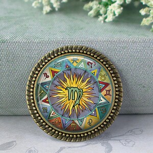 May include: A round, bronze-colored Virgo zodiac sign pendant. The pendant features a colorful design with the Virgo symbol at the center, surrounded by other zodiac symbols and a sunburst pattern. The pendant has a detailed, textured border.