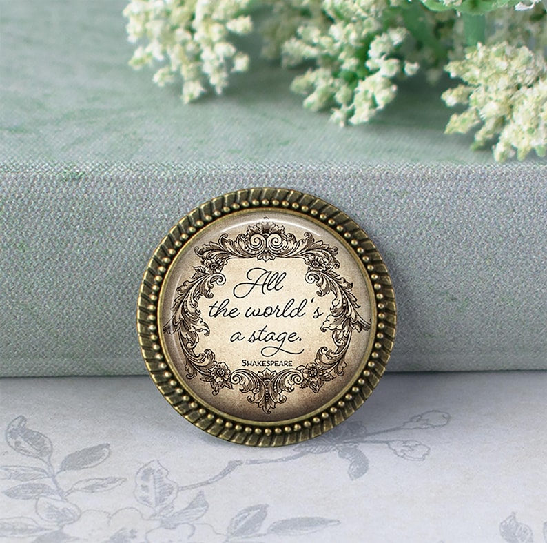 May include: A round, antique-style pin with the quote "All the world's a stage" by Shakespeare. The pin has a bronze-colored, beaded edge and a cream-colored center with ornate floral and scroll designs. The text is in a classic, elegant font.