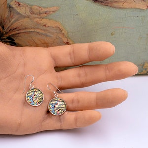 May include: A pair of silver-tone dangle earrings. Each earring features a circular design with a map-like pattern in shades of yellow, blue, and red, with text that reads "Clemson" and other place names. The earrings are held in a hand.