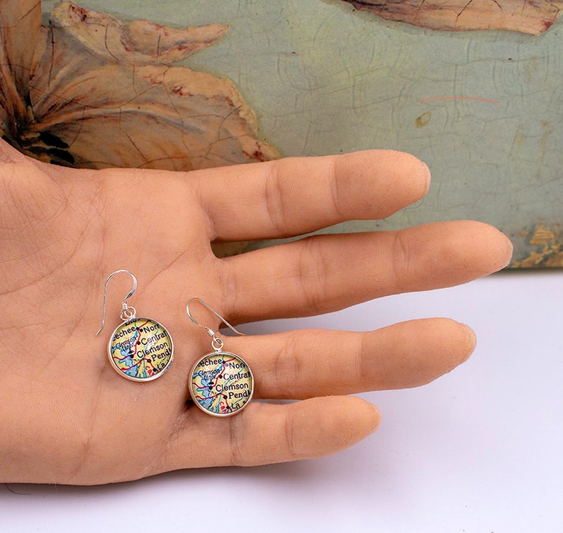 May include: A pair of silver-tone dangle earrings. Each earring features a circular design with a map-like pattern in shades of yellow, blue, and red, with text that reads "Clemson" and other place names. The earrings are held in a hand.
