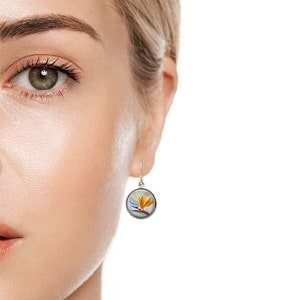 May include: A silver-toned, round earring with a hook closure. The earring features a colorful illustration of a bird of paradise flower in shades of orange, purple, and blue. The earring is shown on a person's ear.