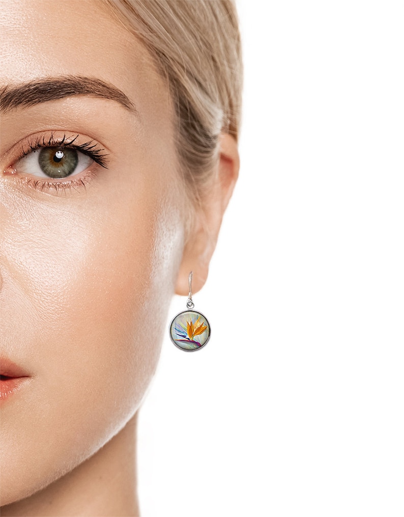 May include: A silver-toned, round earring with a hook closure. The earring features a colorful illustration of a bird of paradise flower in shades of orange, purple, and blue. The earring is shown on a person's ear.