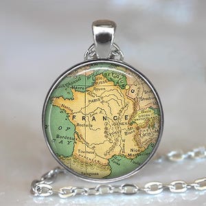 K&ouml;nnte beinhalten: Eine silberfarbene Halskette mit Anh&auml;nger, die einen kreisf&ouml;rmigen Anh&auml;nger mit einer Karte von Frankreich zeigt. Die Karte ist in Gr&uuml;n- und Beiget&ouml;nen gehalten, mit dem Wort "FRANCE" deutlich sichtbar. Die Halskette hat eine feine Kette.