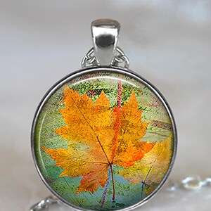 May include: A silver pendant necklace with a round glass charm featuring a yellow and orange maple leaf on a green and blue background.