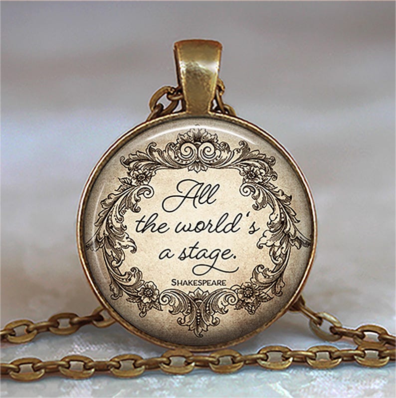 May include: A round, antique gold-toned pendant necklace. The pendant features the quote "All the world's a stage" by Shakespeare, surrounded by an ornate floral design. The chain is also gold-toned.