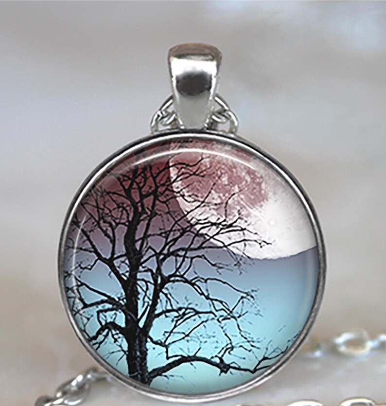K&ouml;nnte beinhalten: Eine silberne Halskette mit einem runden Glasanh&auml;nger, der eine schwarze Silhouette eines Baumes vor dem Hintergrund eines roten und blauen Mondes zeigt.