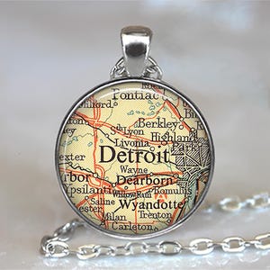 May include: A silver-toned pendant necklace featuring a circular pendant with a map of Detroit, Michigan. The map is in shades of yellow, red, and black, with the city name prominently displayed. The necklace chain is also silver.