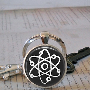 May include: A silver keychain with a round pendant featuring a white atom symbol on a black background. The keychain is attached to a silver key ring. A key is visible in the background.