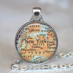 May include: A round silver pendant featuring a map of New York City. The map shows the boroughs of Bronx, Brooklyn, Queens, and Manhattan, with the city name in bold letters. The pendant hangs from a silver chain.