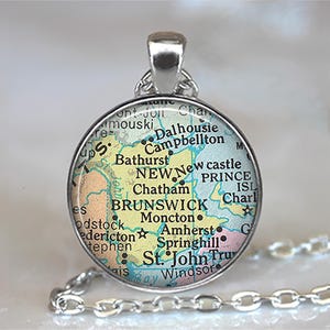 May include: A silver-toned pendant necklace featuring a circular charm with a map design. The map highlights the New Brunswick region with place names like Moncton and Fredericton. The chain is a delicate link style.