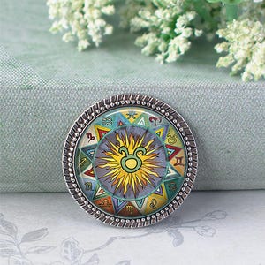 Op de afbeelding: Een ronde, zilverkleurige hanger met een kleurrijk dierenriemdesign. De centrale afbeelding is een zon met het Stier-symbool, omringd door andere astrologische symbolen. De hanger heeft een kralenrand.