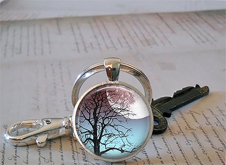K&ouml;nnte beinhalten: Ein silberner Schl&uuml;sselanh&auml;nger mit einem runden Glasanh&auml;nger, der eine schwarze Silhouette eines Baumes vor einem blauen und rosa Himmel zeigt. Der Anh&auml;nger ist an einem silbernen geteilten Ring und einem silbernen Karabinerhaken befestigt.