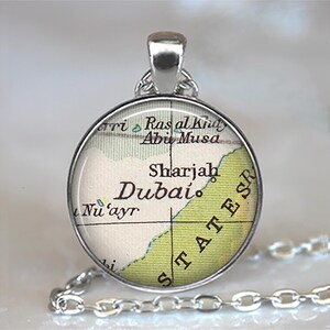 May include: Silver-toned pendant necklace featuring a round charm with a map of Dubai and Sharjah. The map has green and beige tones with black text. The chain is a delicate silver link.