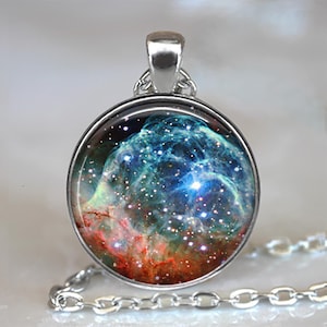 May include: A silver-toned pendant necklace featuring a round, glass-covered charm. The charm displays a vibrant nebula design with shades of blue, red, and white, and a silver chain. The necklace is a celestial-themed accessory.