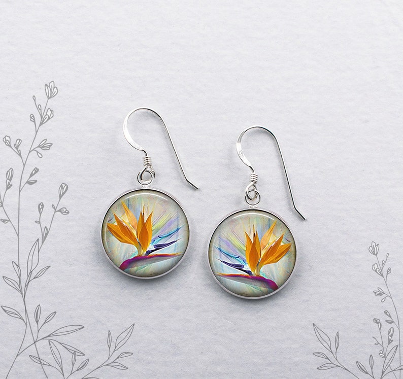 May include: Silver dangle earrings with round, silver-rimmed, glass cabochons featuring a colorful bird of paradise flower illustration on a white background.