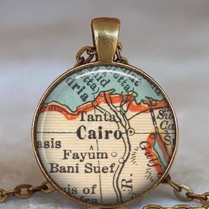 May include: A round pendant necklace with a map of Cairo, Egypt. The pendant has a gold-colored frame and chain. The map is in shades of blue, orange, and beige, with the city name "Cairo" prominently displayed.