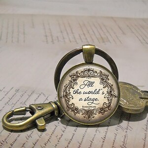 May include: A vintage-style keychain with a round pendant featuring the quote "All the world's a stage" by Shakespeare. The pendant has a decorative border and is attached to a bronze-colored clasp and key ring. The background shows handwritten script.