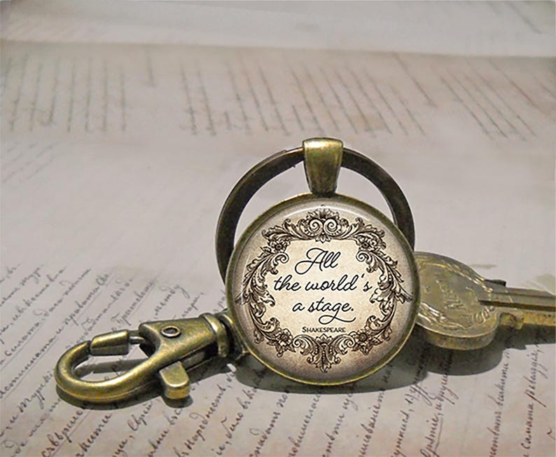 May include: A vintage-style keychain with a round pendant featuring the quote "All the world's a stage" by Shakespeare. The pendant has a decorative border and is attached to a bronze-colored clasp and key ring. The background shows handwritten script.