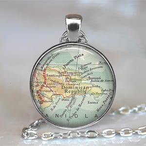May include: A silver-toned pendant necklace featuring a round glass charm with a map of the Dominican Republic. The map is in shades of green, red, and beige, with place names visible. The chain is a delicate silver link.