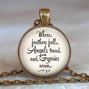 May include: A round pendant necklace with a gold-colored chain. The pendant has a gold-colored frame and a white background with the text "Where feathers fall, Angels tread and Gypsies roam" in black script.