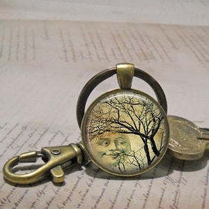 May include: A round bronze keychain featuring a glass cabochon with a graphic print of a human face partially obscured by the dark, bare branches of a tree against a pale yellow background. The keychain includes a metal lobster clasp and ring.
