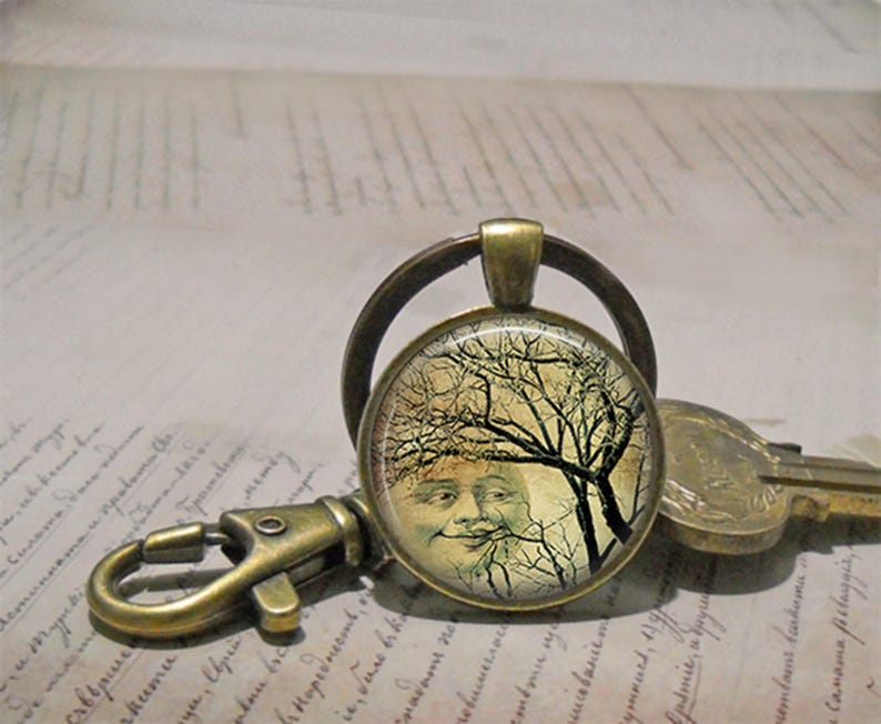 May include: A round bronze keychain featuring a glass cabochon with a graphic print of a human face partially obscured by the dark, bare branches of a tree against a pale yellow background. The keychain includes a metal lobster clasp and ring.