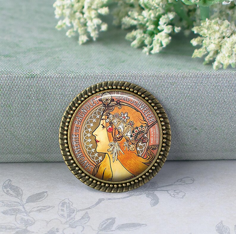 May include: Round bronze-colored pin featuring an Art Nouveau style illustration of a woman with flowing auburn hair and ornate headwear. The pin has a detailed border and includes the text "SAVONNERIE DE BAGNOLET" and "ALFRED SCHWEIZER".