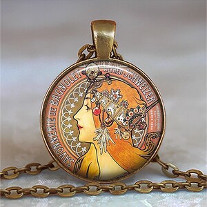 May include: A round pendant necklace with a vintage Art Nouveau design. The pendant features a profile of a woman with red hair and a decorative headpiece. The pendant is gold-toned and hangs from a matching chain.