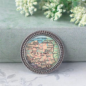 May include: A round, silver-toned pin featuring a map of Poland. The map is in shades of red, yellow, and green, with cities labeled. The pin has a textured, beaded border. The background is a soft, muted green.