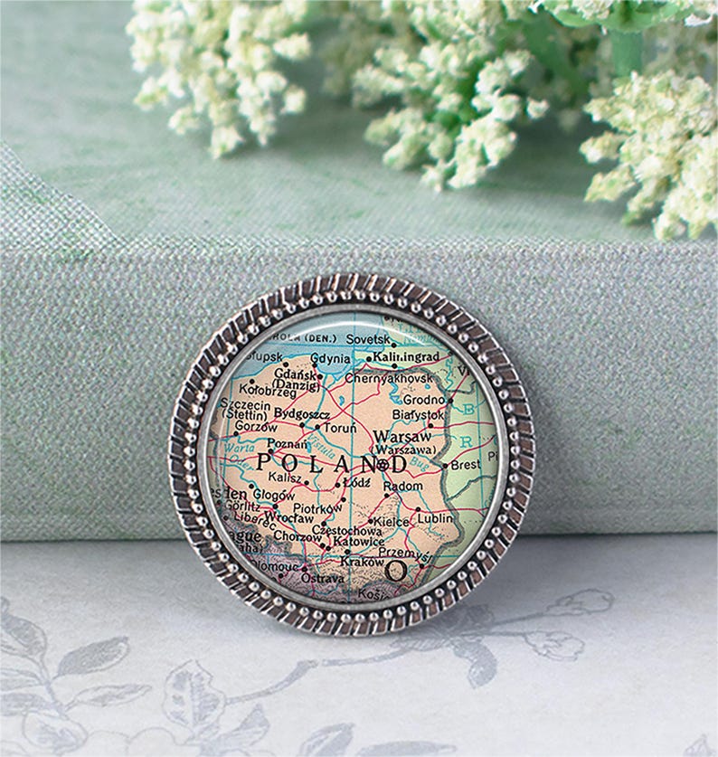 May include: A round, silver-toned pin featuring a map of Poland. The map is in shades of red, yellow, and green, with cities labeled. The pin has a textured, beaded border. The background is a soft, muted green.
