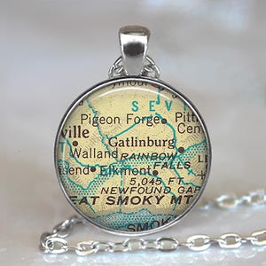 May include: A round silver pendant with a map of Gatlinburg, Tennessee, featuring the text "Gatlinburg" and "Smoky Mt." The pendant hangs from a silver chain. The map has a yellow background with blue and brown details.
