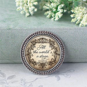 May include: A round, silver-toned pin with a decorative border. The pin features the quote "All the world's a stage" in elegant script, surrounded by ornate floral and scrollwork. The Shakespeare quote is centered on a cream-colored background.