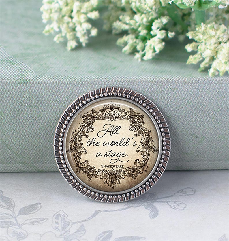 May include: A round, silver-toned pin with a decorative border. The pin features the quote "All the world's a stage" in elegant script, surrounded by ornate floral and scrollwork. The Shakespeare quote is centered on a cream-colored background.