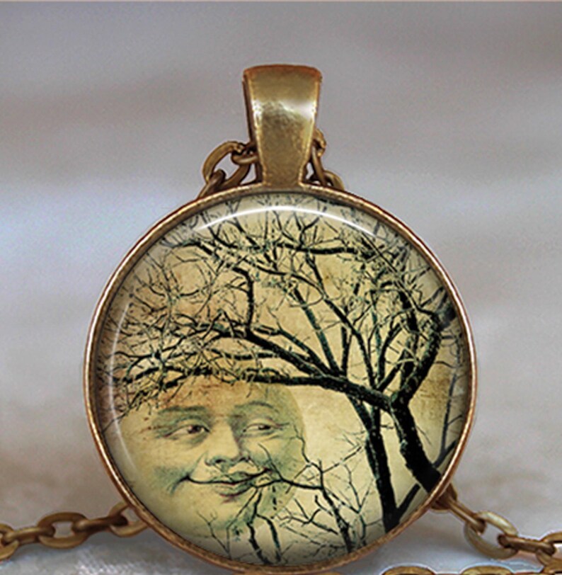 May include: A round pendant necklace featuring a bronze-toned metal frame and a glass cabochon. The image inside depicts a vintage-style illustration of a smiling moon face partially obscured by the dark, intricate branches of a bare tree.