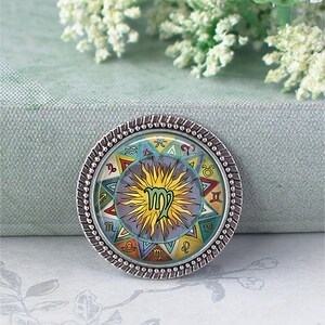 May include: A round, silver-toned pendant featuring a colorful zodiac design. The center displays the Virgo symbol, surrounded by other astrological symbols and a sunburst design. The pendant has a textured, beaded edge.