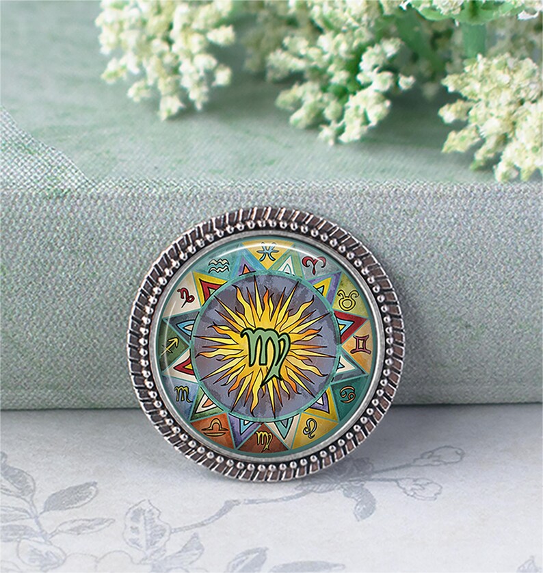 May include: A round, silver-toned pendant featuring a colorful zodiac design. The center displays the Virgo symbol, surrounded by other astrological symbols and a sunburst design. The pendant has a textured, beaded edge.
