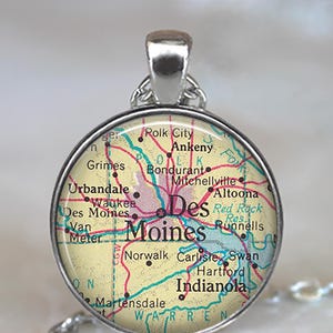 May include: A round pendant with a map of Des Moines, Iowa, featuring the city name in a bold font. The map includes surrounding towns and roads in red and blue, set against a cream background. The pendant has a silver-colored metal frame and bail.