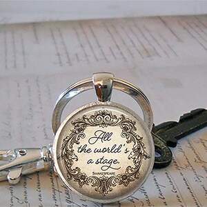 May include: Silver keychain with a round pendant featuring the quote "All the world's a stage" in elegant script, surrounded by a decorative floral border. A key and clasp are also visible.