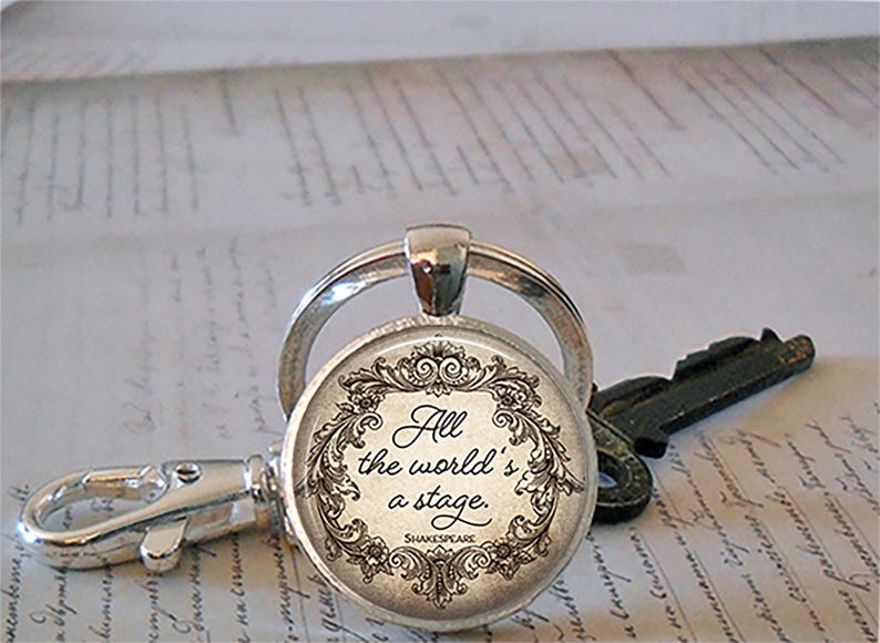 May include: Silver keychain with a round pendant featuring the quote "All the world's a stage" in elegant script, surrounded by a decorative floral border. A key and clasp are also visible.
