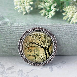 May include: A round metal button featuring a vintage-style illustration of a smiling moon face partially obscured by dark, bare tree branches. The button has a textured, silver-toned metallic rim with a beaded edge surrounding the central image.