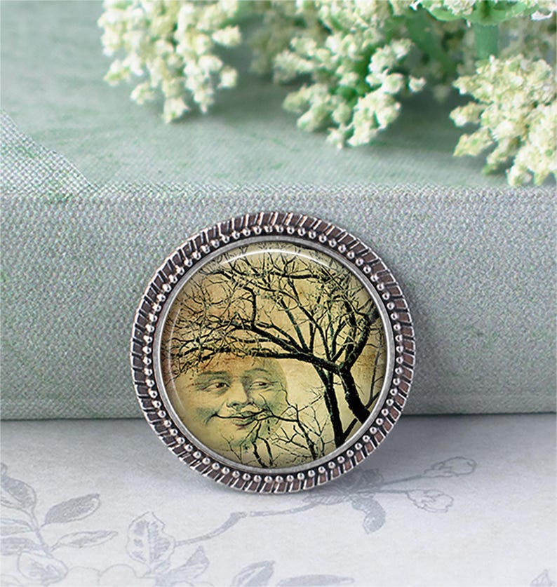 May include: A round metal button featuring a vintage-style illustration of a smiling moon face partially obscured by dark, bare tree branches. The button has a textured, silver-toned metallic rim with a beaded edge surrounding the central image.