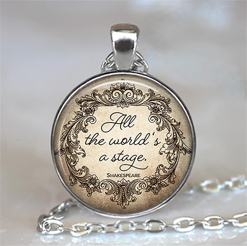 May include: Silver-toned pendant necklace featuring a round charm with the quote "All the world's a stage" in elegant script. The quote is framed by ornate floral and scrollwork designs, with the name "Shakespeare" below. The chain is a delicate silver link.