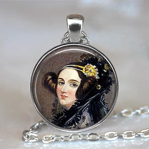 May include: A round silver pendant featuring a portrait of a woman with dark hair and a black lace shawl. The portrait is set against a muted background, and the pendant hangs from a silver chain.