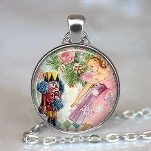 May include: Silver-toned pendant necklace featuring a round charm with a vintage illustration. The charm depicts a nutcracker soldier, a ballerina in a pink dress, and winter elements. The background includes pink roses and evergreen branches.