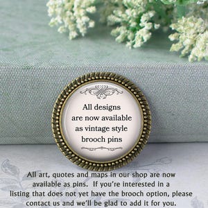 a decorative pin with text, placed on a surface with a floral pattern. The pin announces that all designs are now available as vintage style brooch pins, and that all art, quotes, and maps in the shop are now available as pins. If someone is interested in a listing that does not yet have the brooch option, they should contact the shop and the staff will be happy to add it for them.
